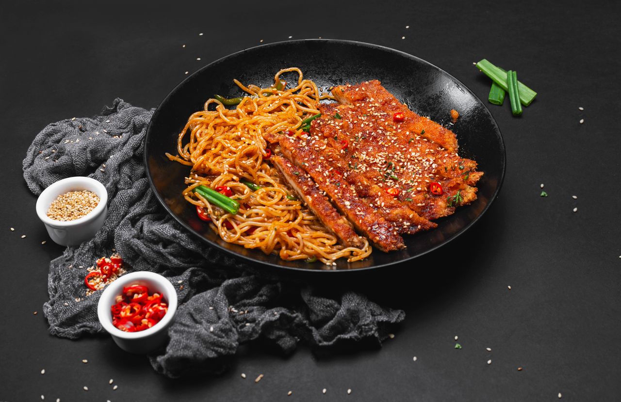Delicious spicy grilled chicken and noodles garnished with sesame on a sleek black plate.